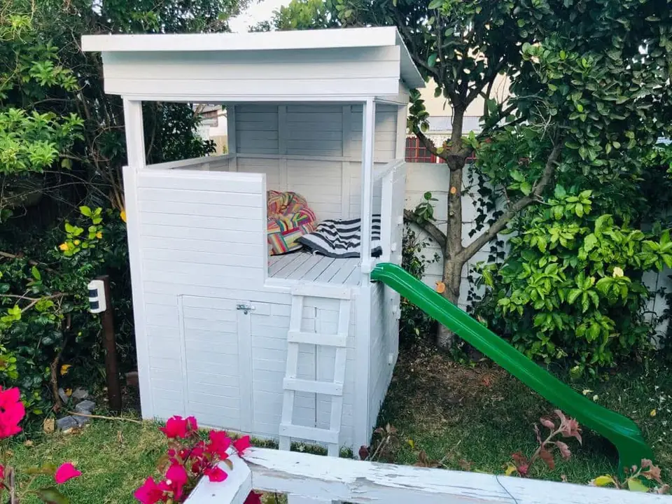 White painted timber jungle gym with a covered platform, ladder, storage door and green slide in a garden